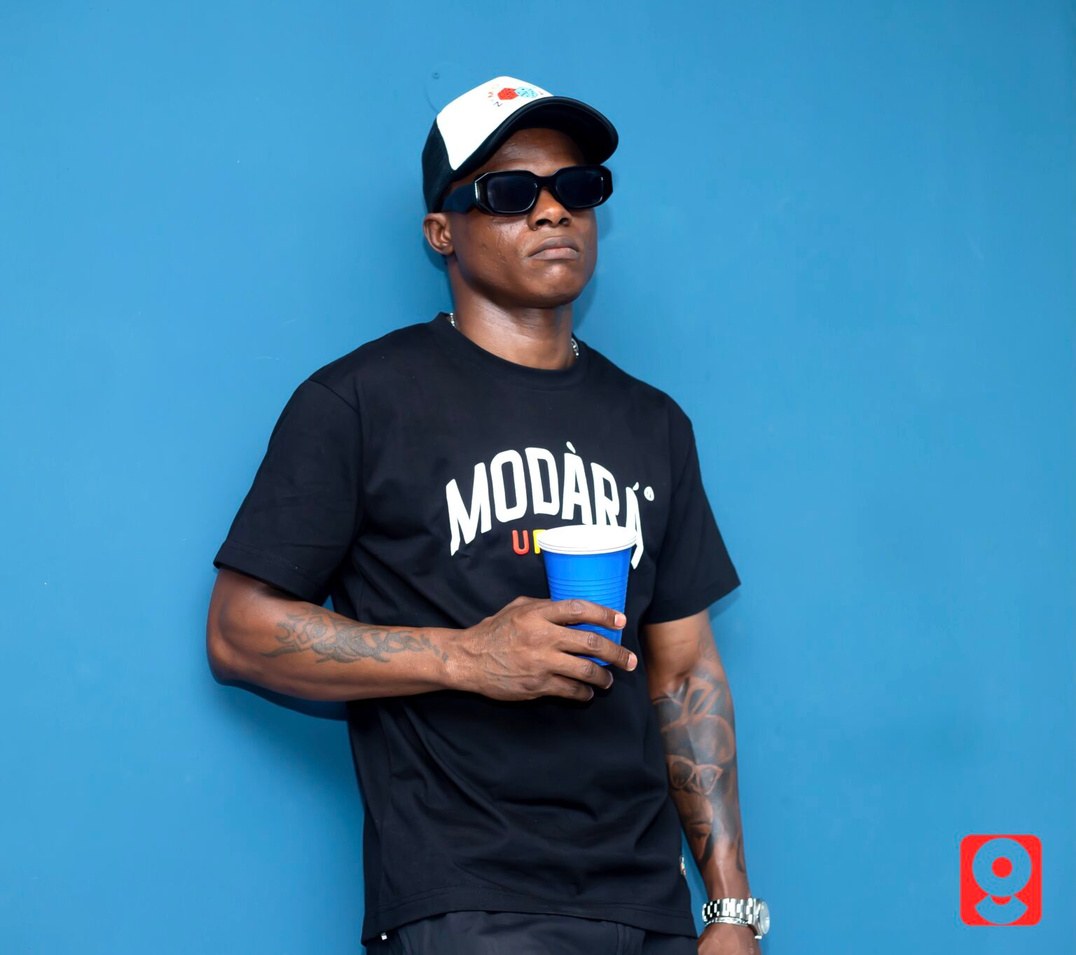 DJ Kenny in a Modari tee holding a cup against a plain wall